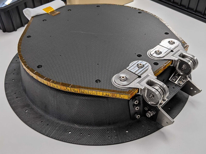 Image shows the Psyche optics enclosure with its attached deployable cover placed on a flat grey surface. It is a composite/metallic hybrid structure. The primary structure is made of carbon fiber with metallic hinge component.  It is a round structure with a wide flat edge around the bottom that has attachment holes. The deployable cover is flat . The lid is protected along the opening edge with a folded white tissue.